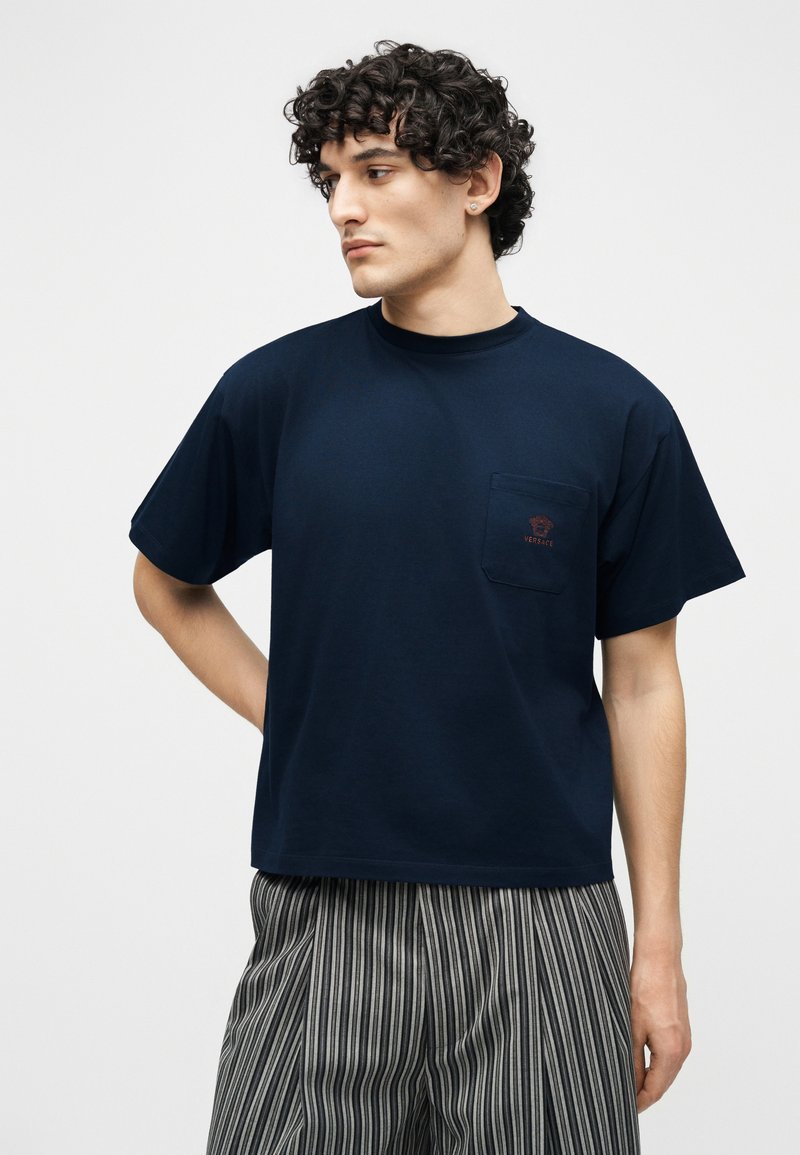 Giovane uomo con capelli ricci che indossa una maglietta blu navy con un piccolo logo sulla tasca e pantaloni a righe verticali bianche e nere, guarda a sinistra.