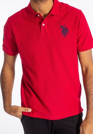 Mężczyzna w czerwonej koszulce polo z krótkim rękawem, z ciemnoniebieskim haftowanym logo gracza polo na piersi, ręką w kieszeni, na białym tle.