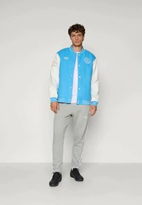 Blue and white varsity jacket with embroidered logo and patches, worn over a white T-shirt and grey sweatpants. Black athletic shoes.