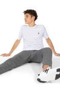 Bílá košile s krátkým rukávem a texturovaným vzorem, doplněná o černé logo pavouka, v kombinaci se šedými zúženými kalhotami a bílými sportovními botami.