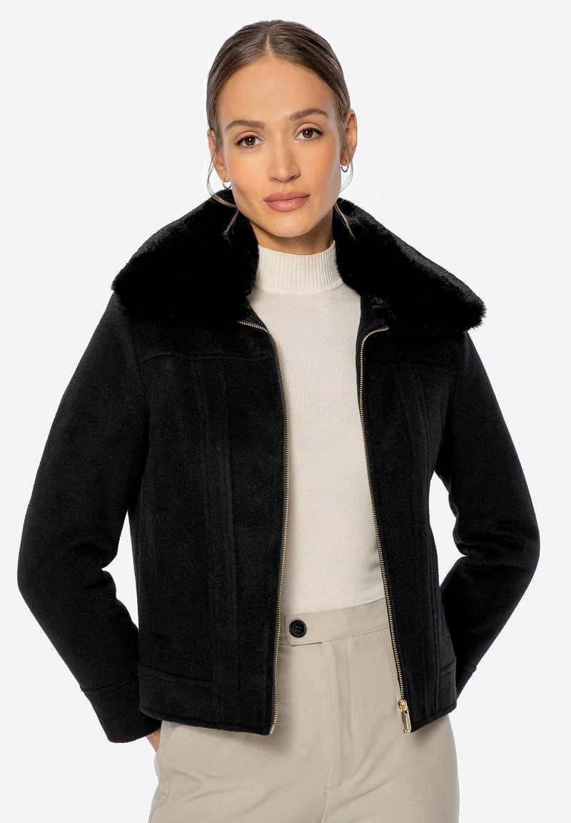 Chaqueta negra con cuello de felpa, hecha de tela suave. Cuenta con una cremallera frontal, detalles de costura vertical y un corte corto.