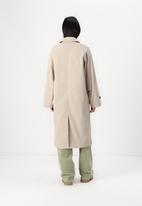 Manteau beige avec une coupe décontractée, manches longues et fente au dos, porté sur un pantalon vert clair. Le tissu semble lisse avec un design structuré.
