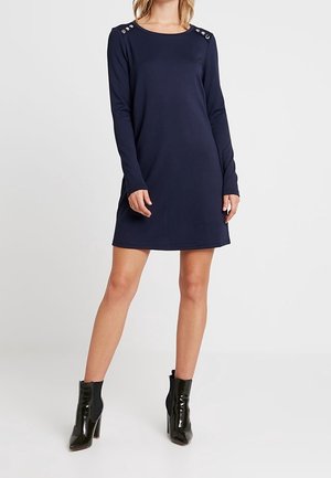 Donna che indossa un abito lungo a maniche lunghe di colore blu navy con dettagli a bottoni sulle spalle e stivali neri in vernice alla caviglia, in piedi su uno sfondo semplice.