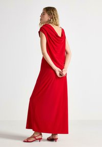 Abito maxi senza maniche di colore rosso con schiena drappeggiata. Il tessuto è liscio e fluido, abbinato a tacchi a spillo rossi coordinati.