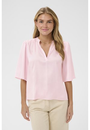 Pika blondide juustega naine, kes kannab heleroosat, küünarnukipikkuste varrukatega pluusi ja beeže pükse, naeratades kaamerale.