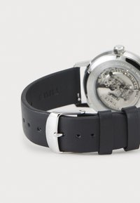 Black leather watch strap with silver buckle engraved "TIMEX," attached to a stainless steel watch showing a see-through back with internal mechanics.