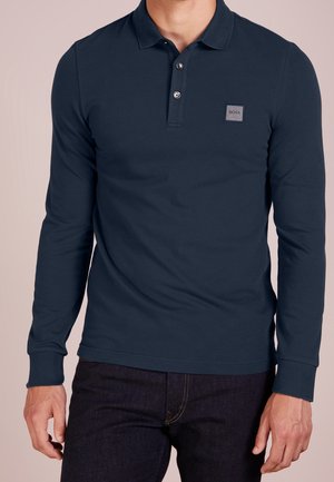 Mężczyzna ubrany w granatową, długorękawową koszulkę polo z guzikami i małym kwadratowym logo "BOSS" na piersi, w połączeniu z ciemnymi dżinsami.