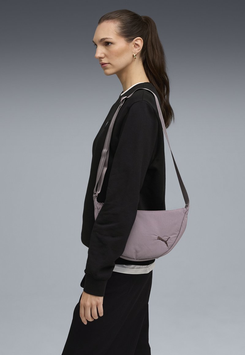 Jasnofioletowa torebka typu crossbody o gładkiej fakturze, z wyraźnym logo, regulowanym paskiem oraz eleganckim, dopasowanym kształtem.