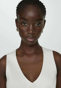 Haut sans manches de couleur claire avec un décolleté en V, présentant un tissu texturé et un détail de bouton central. Le modèle a les cheveux tressés et porte des boucles d'oreilles.
