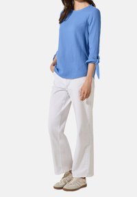 Haut bleu texturé à manches retroussées, associé à un pantalon blanc à jambes larges et des baskets beiges avec des accents marron.