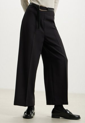 Personne portant un pantalon noir à jambes larges, une ceinture noire, des mocassins noirs et un haut de couleur claire, debout sur un fond de sol et de mur clairs et unis.
