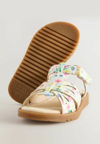 Blommönstrade sandaler med justerbara remmar, mjuk fotbädd och en strukturerad gummisula. Färgerna inkluderar pastellblommor på vit bakgrund.