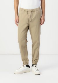 Pantalones jogger beige con cordón y puños elásticos, fabricados en una tela suave, combinados con zapatillas blancas con un acento negro en el talón.