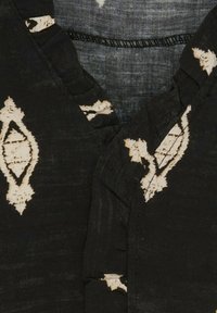 Sort stof top med V-halsdesign, prydet med hvide diamantmønstre og teksturerede flæseaccentuer langs udskæringen.