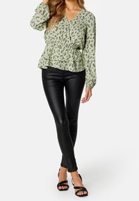Blouse peplum floral verte avec des manches longues, décolleté en V et ourlet à volants, associée à un pantalon noir ajusté en simili cuir et des sandales à talons noires.