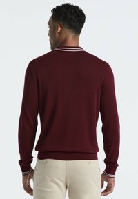 Bordeauxfarbener Pullover mit strukturiertem Strickmuster, ausgestattet mit einem weiß gestreiften Kragen und Bündchen sowie einer figurbetonten Passform, getragen über hellen Hosen.