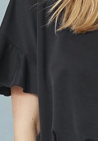 Zwarte top met gerimpelde mouwen, gemaakt van een gladde stof. Het ontwerp is eenvoudig en ontspannen, met nadruk op comfort en bewegingsvrijheid.