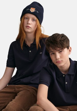Polo-shirts navy en coton, avec une patte de boutonnage à deux boutons et un logo brodé ; portés avec un pantalon en velours côtelé brun.