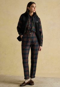 Quiltowana czarna kamizelka na koszuli w kratę i dopasowanych ciemnoniebieskich spodniach z czerwonymi i zielonymi akcentami. Brązowe mokasyny z wzorzystymi skarpetkami.