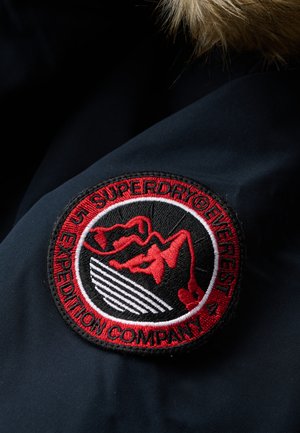 Rund patch med bjergdesign, rød og hvid broderi på sort baggrund, med teksten "SUPERDRY® EXPEDITION COMPANY".