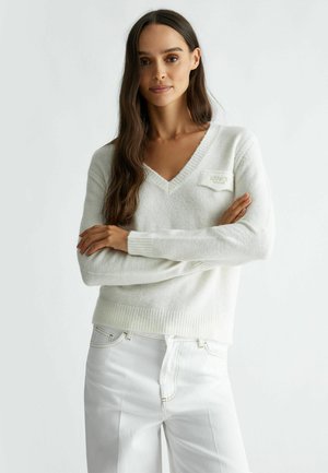 Maglione bianco a maglia con scollatura a V, orlo a coste e un piccolo etichetta. Abbinato a pantaloni bianchi, che mostrano una consistenza morbida e una vestibilità comoda.