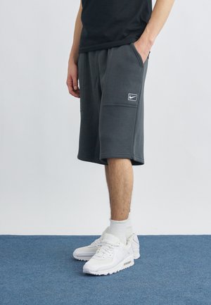 Άντρας που φοράει σκούρα γκρι σορτς Nike και λευκά αθλητικά παπούτσια Nike, στεκόμενος σε μπλε χαλί μπροστά σε απλό γκρι ανοιχτό φόντο.