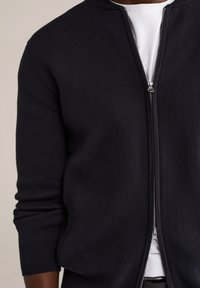 Tamsiai mėlynas megztinis su užtrauktuku, turintis ribotuotą tekstūrą, suapvalintu kaklu ir šoninėmis kišenėmis. Priekinė dalis su subtiliu sidabriniu užtrauktuku.