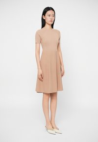 Vestido de malha bege canelado com corpete ajustado, mangas curtas e saia evasê. Emparelhado com sapatos brancos de bico fino.