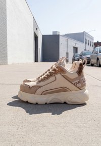 Par beige og brune chunky sneakers placeret på en urban fortov med biler og bygninger i baggrunden.
