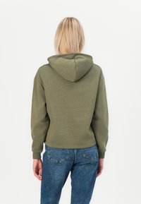 Olíva zöld kapucnis pulóver, laza fitnesz, bordás mandzsettákkal és aljjal, puha tapintású anyaggal. Kék farmernadrággal párosítva.