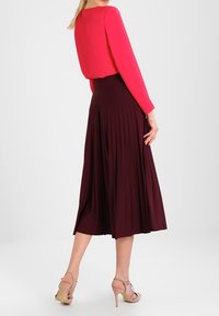 Rode top met lange mouwen gecombineerd met een vloeiende bordeauxrode midi-jurk. Materiaal lijkt zacht; de jurk heeft plooien en een hoge taille.