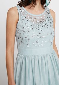 Svetle modré šaty s přiléhavým korzetem, který je zdobený květinovou výšivkou a flitry. Rozšířená sukně má hladkou texturu a průhledný výstřih.