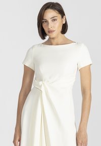 Kremowa sukienka z krótkimi rękawami, dekoltem w łódkę, w detalach węzła w talii i miękkimi fałdami opadającymi z talii. Gładka tekstura materiału.
