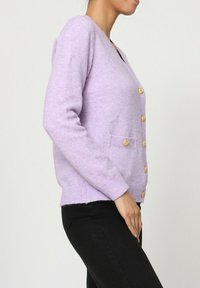 Cardigan lilas en tissu doux, avec un col en V, des manches longues, des boutons dorés et deux poches avant. Ourlet et poignets simples à côtes.