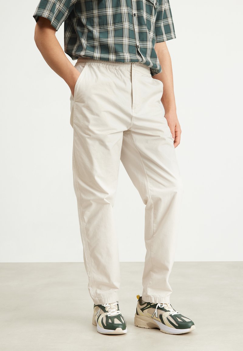 Pantalons léger beige, à taille élastique, avec poches latérales, légère brillance et coupe décontractée, associés à des baskets de sport vertes et beiges.