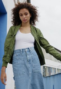 Zelená bomber bunda, bílý top bez rukávů a modrá deninová sukně s náplety. Modelka stojí na pozadí v modro-bílém stylu.