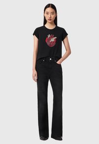 Γυναίκα με μακριά σκούρα μαλλιά που φοράει μαύρα τζιν και μαύρο t-shirt με σχέδιο ανατομικής καρδιάς σε κόκκινο και λευκό.