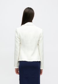 Blazer blanc texturé avec ourlet et col effilochés, manches longues. Porté sur une jupe bleu foncé, affichant un design simple et ajusté.