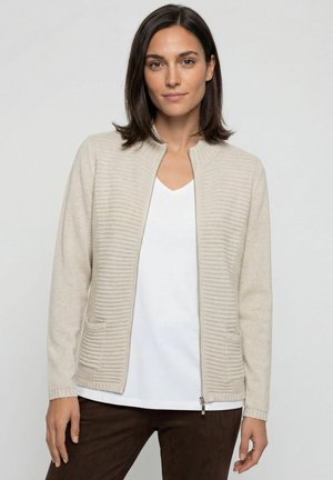 Vrouw met donker haar tot op de schouders, die een beige cardigan met rits draagt over een wit overhemd en donkerbruine broek, staand voor een effen lichte achtergrond.