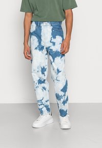 Blue and white tie-dye jeans with a relaxed fit, featuring a zipper fly, five pockets, and contrasting hardware, paired with white sneakers.