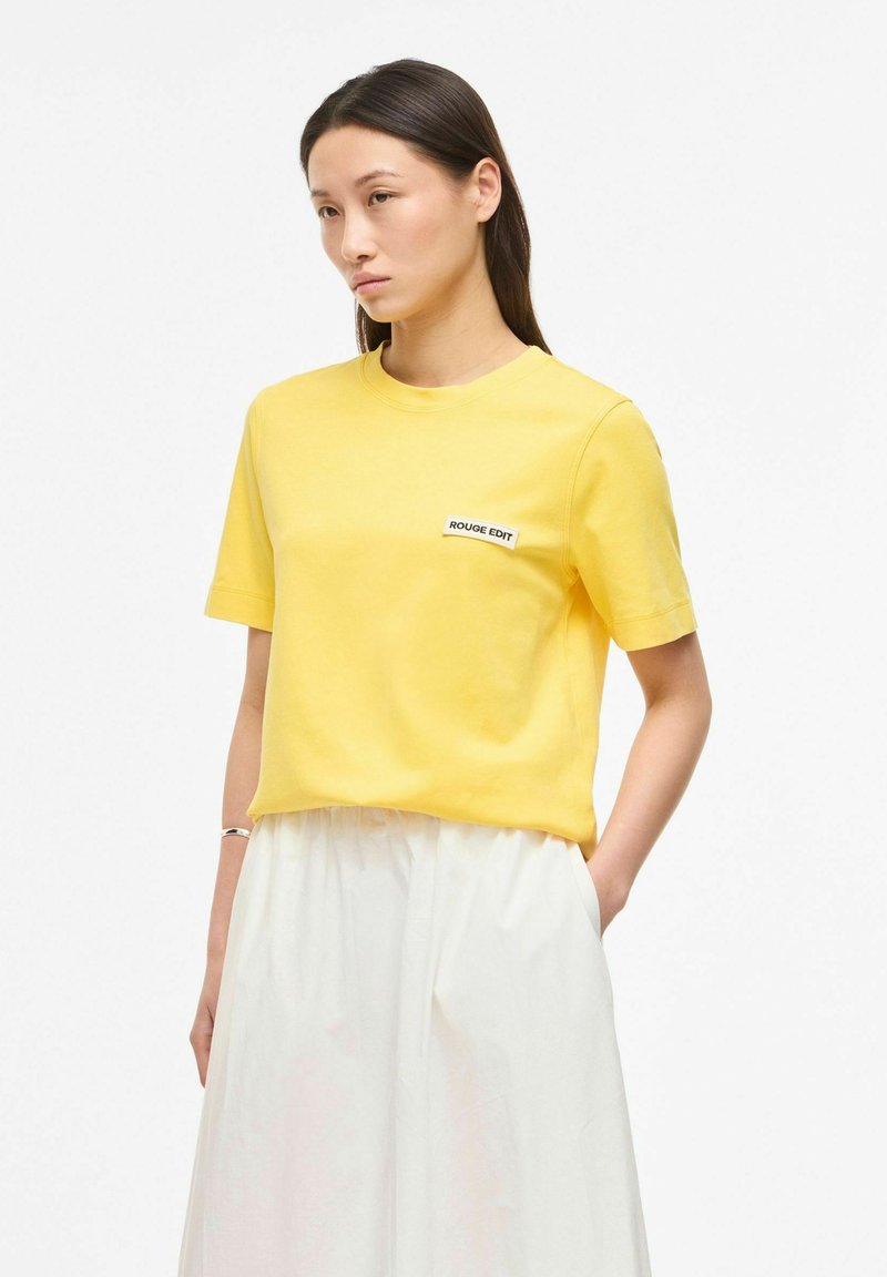 Femme aux cheveux raides et foncés portant un t-shirt jaune à manches courtes avec un écusson "ROUGE EDIT" et un pantalon blanc taille haute, les mains dans les poches.