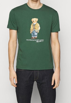 T-shirt vert avec un ours en peluche portant une veste, un pull, un jean et des chaussures, avec le texte "Polo Bear by Ralph Lauren" en dessous.
