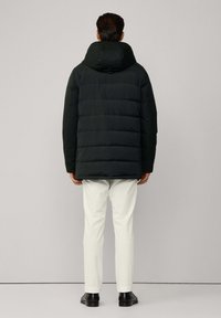 Muškarac stoji okrenut leđima, obučen u crnu puffer jaknu s kapuljačom, bijele hlače i crne cipele, nasuprot jednostavnoj sivoj pozadini.