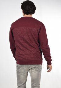 Bordeauxfarbener Crewneck-Sweatshirt mit strukturiertem Rückenteil. Weiches Baumwollmaterial. Enganliegende Bündchen und Taillenbund. Getragen mit hellgrauen Jeans.
