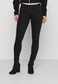 Mujer vestida con vaqueros negros ajustados y botines de tacón negros de tobillo, de pie contra un fondo liso de color gris claro.