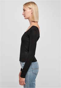 Črn rebrast dolg rokav s off-shoulder dizajnom, prilegajoča silhueta in mehak material, v kombinaciji s svetlo modrimi oprijetimi kavbojkami.