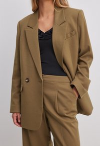 Oliwkowa marynarka dopasowana, z ostrymi klapami, pojedynczym zapięciem na guzik i kieszenią z przodu. Nosić na czarnej bluzce, zaprezentowana w luźny sposób.
