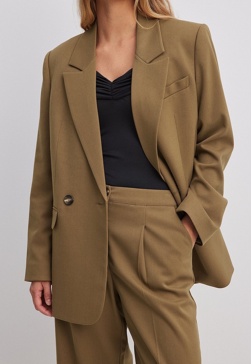 Oliwkowa marynarka dopasowana, z ostrymi klapami, pojedynczym zapięciem na guzik i kieszenią z przodu. Nosić na czarnej bluzce, zaprezentowana w luźny sposób.