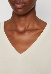 T-shirt con scollo a V di colore crema chiaro, realizzato in tessuto morbido. Presenta una texture liscia e un colletto pulito, con un'attenzione particolare alla zona superiore del torace.