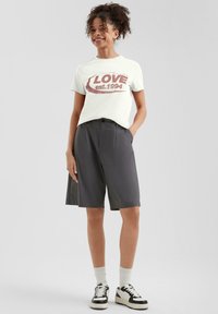 Cremefarbenes T-Shirt mit rotem "LOVE est. 1994" Grafik, kombiniert mit dunklen Shorts. Das Model trägt weiße Socken und schwarze Sneakers mit cremefarbenen Akzenten.